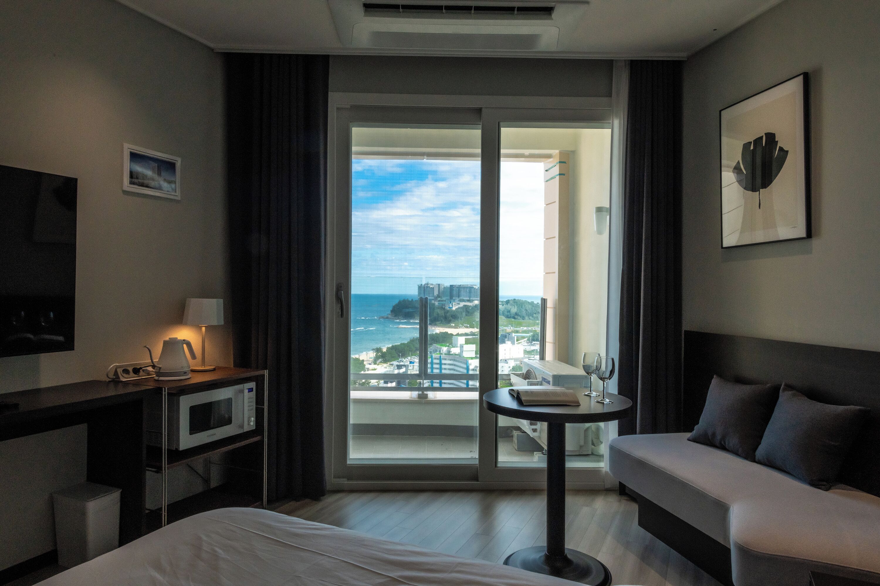 Quarto de hotel com cama, escrivaninha com micro-ondas, pequena mesa com uma taça de vinho, sofá e grande janela com vista para a cidade e mar.