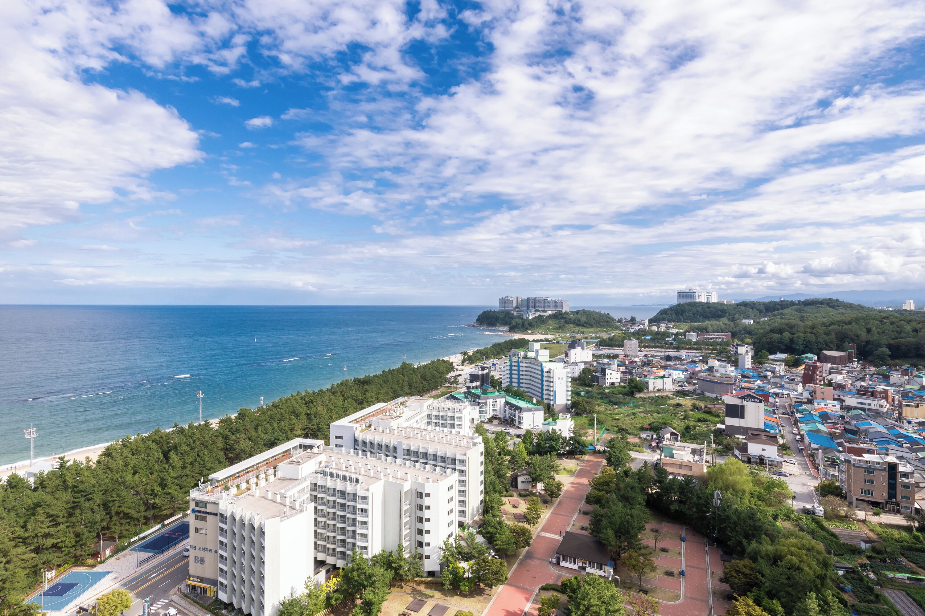 Un paisaje urbano costero con un edificio blanco destacado, una cancha deportiva y vistas al mar.