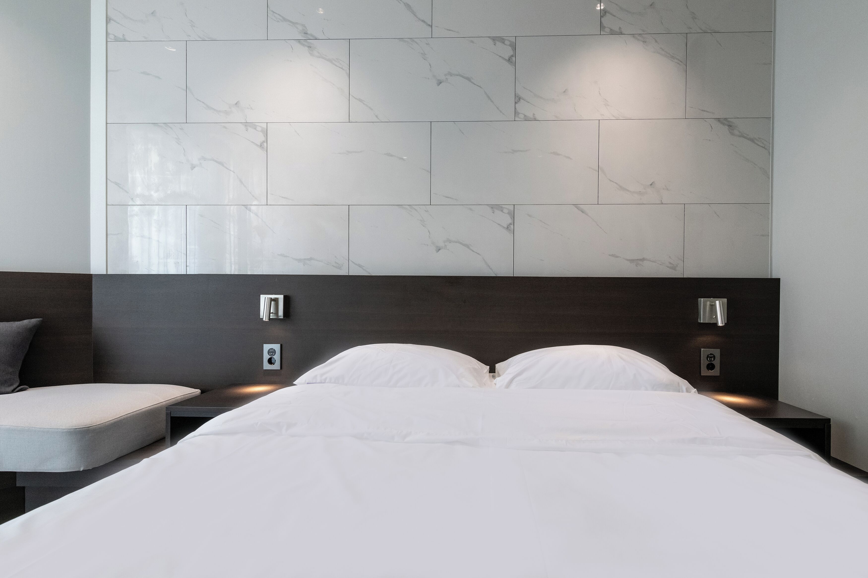 A modern hotel room with a large bed, two bedside tables, a dark headboard, and a marble-patterned wall.