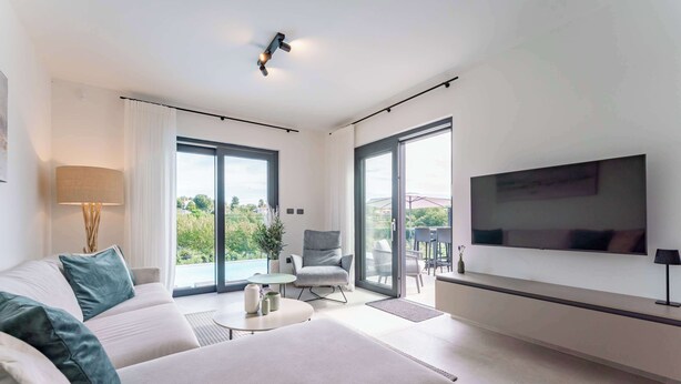 Ein modernes Wohnzimmer mit einer Couch, einem Sessel, einem an der Wand montierten Fernseher und großen Fenstern mit Blick ins Freie.