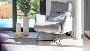 Ein modernes Wohnzimmer mit einem grauen Sessel, einer weißen Vase mit Pflanzen und Blick auf einen Swimmingpool und Grünflächen.