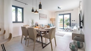 Ein moderner Essbereich mit einem runden Tisch, Stühlen und Pendelleuchten. Im Hintergrund befindet sich eine Couch und ein an der Wand montierter Fernseher.