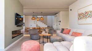 Un salon moderne avec une cheminée, des canapés gris, un pouf rose et un coin repas avec une table et des chaises en bois.