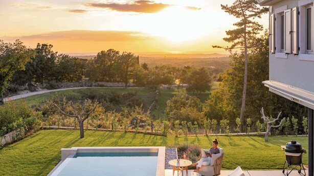 Un espace aménagé au bord de la piscine, offrant une vue sur un jardin paysager et sur des collines lointaines au coucher du soleil.