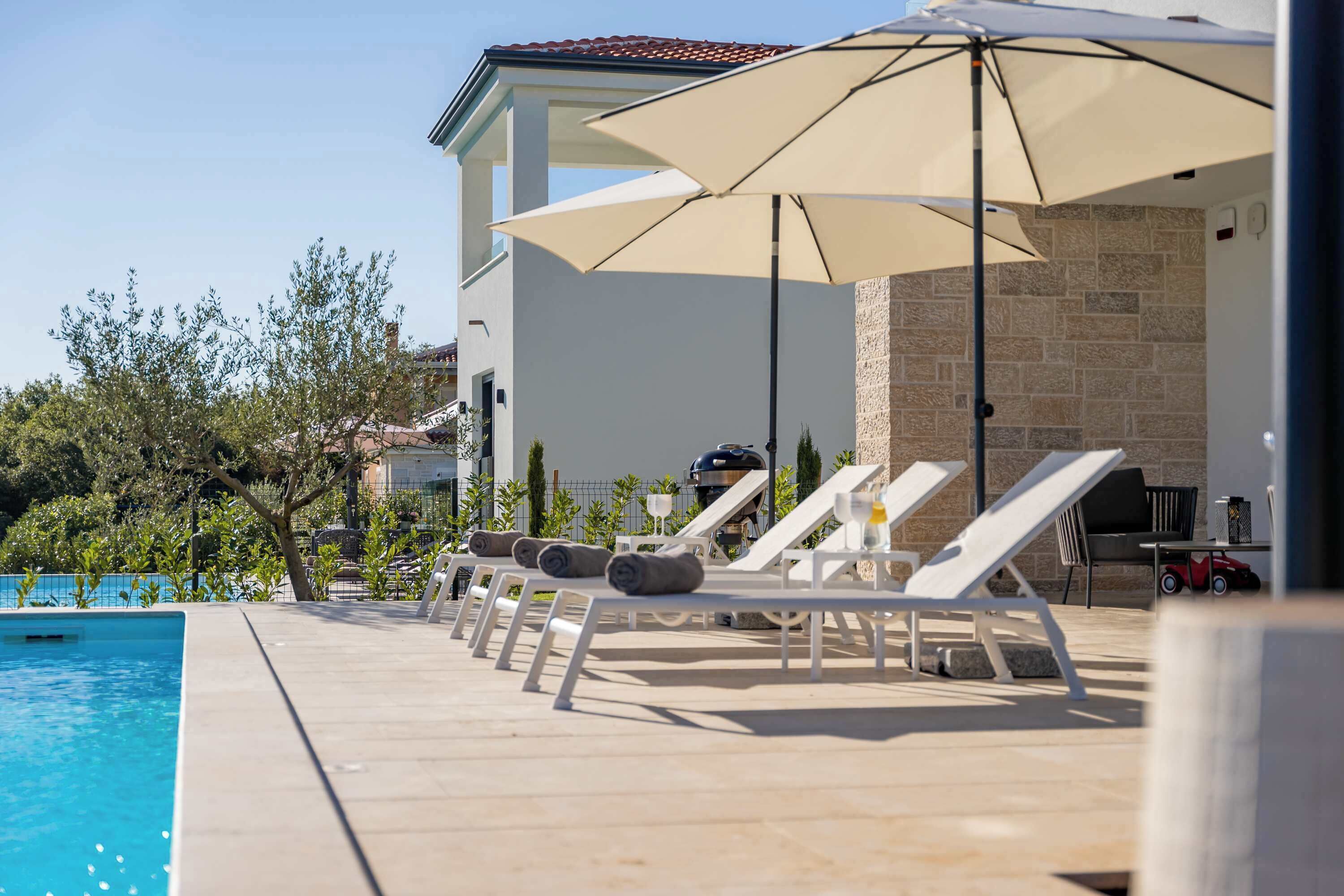 Un espace aménagé au bord de la piscine, avec des chaises longues et des parasols, un mur en pierre et une piscine à l’eau d’un bleu limpide.