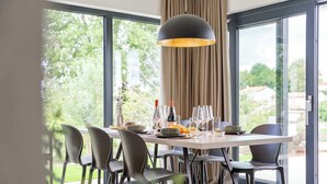 Un coin repas avec une table dressée pour six personnes, entourée de chaises, d’un luminaire suspendu et offrant une vue sur la verdure grâce à de grandes fenêtres.