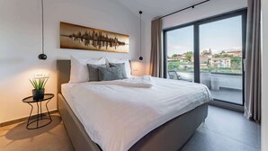 Une chambre moderne avec un grand lit, une vue sur la ville à travers une grande fenêtre, une œuvre d’art représentant des ondes sonores accrochée au mur, et une petite table d’appoint avec une plante.