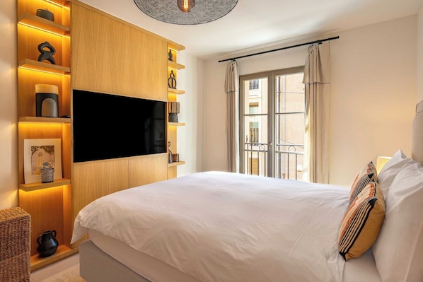 A bedroom with a bed, a TV mounted on the wall, a window with curtains, and a balcony.