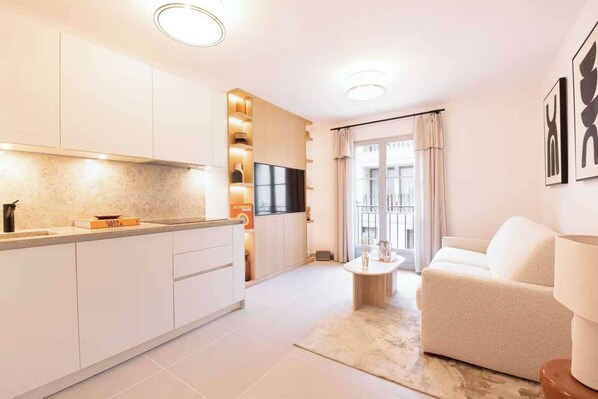 A modern kitchen with white cabinets, a built-in oven, and a flat-screen TV. There is a white sofa and a small round table in the living area.