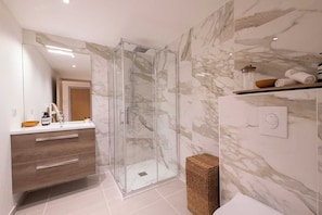 A modern bathroom with a glass shower enclosure, marble walls, a wooden vanity, and a built-in shelf with towels.