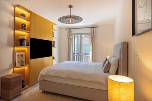 A bedroom with a bed, a TV mounted on a wooden wall, a window with curtains, and a hanging light fixture.
