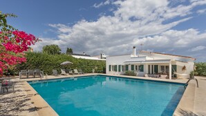 Una piscina con sillas de descanso, un edificio blanco de techo plano y follaje al fondo.
