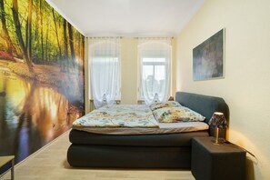 Une chambre à coucher avec un lit, une table de chevet, une lampe, un tableau au mur et une fresque représentant une forêt.