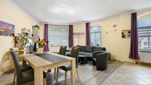 Una sala de estar moderna con comedor, sofá y una mesa de centro. Se ve una ventana con cortinas, un ventilador de techo y una pared decorada con adornos de mariposas.
