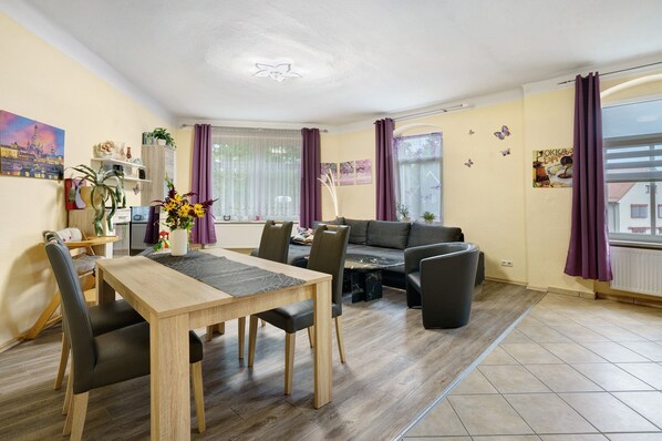Un salon moderne comprenant un coin repas, un canapé et une table basse. On y trouve une fenêtre avec des rideaux, un ventilateur de plafond et un mur décoré de motifs de papillons.