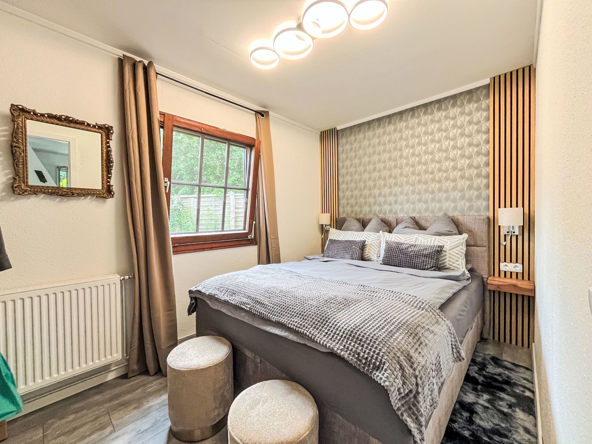 Une chambre à coucher comprenant un lit, deux tabourets ronds, un miroir, une fenêtre avec des rideaux et une lampe fixée au mur.