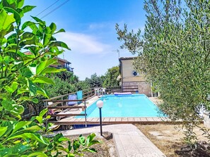 A swimming pool with a wooden deck, surrounded by greenery and a stone wall.