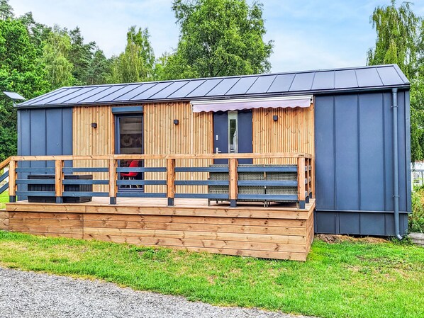 A small wooden cabin with a metal roof, a wooden deck with benches, and a door leading to an interior space.