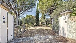 Une allée de gravier mène à une entrée fermée par un portail, elle-même bordée d’un mur de pierres et de grilles en métal.