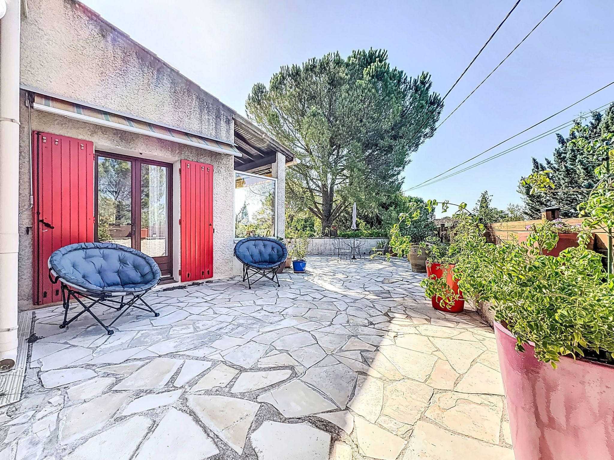 Une terrasse avec un sol en pierre, deux chaises bleues, une porte rouge et des plantes en pot.
