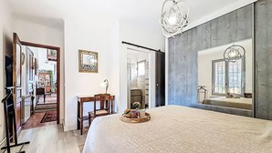Une chambre avec un grand lit, un bureau, une chaise et un miroir. Une porte donne accès à une autre pièce.