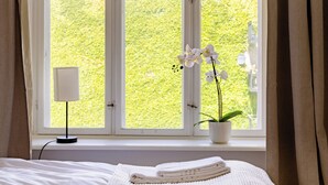 Un dormitorio con un ventanal grande, una lámpara de noche, una planta en maceta y una cama bien hecha.