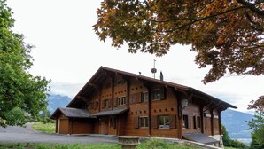 A wooden chalet with a sloped roof, multiple windows, and a stone fountain in the foreground. - Champ de Brent by Interhome (Ollon)