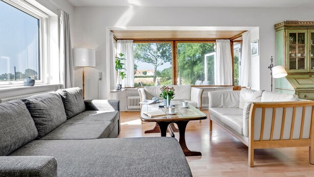 Un salón amplio con un sofá seccional gris, una mesa de centro de madera y un sillón blanco. Cerca de una ventana grande con cortinas, se encuentra una mesa de comedor con sillas.