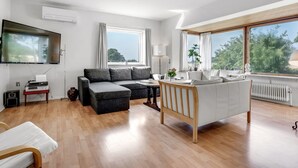 Una sala de estar moderna con un sofá seccional gris, un sillón blanco, una mesa de comedor de madera y un televisor de pantalla plana montado en la pared.