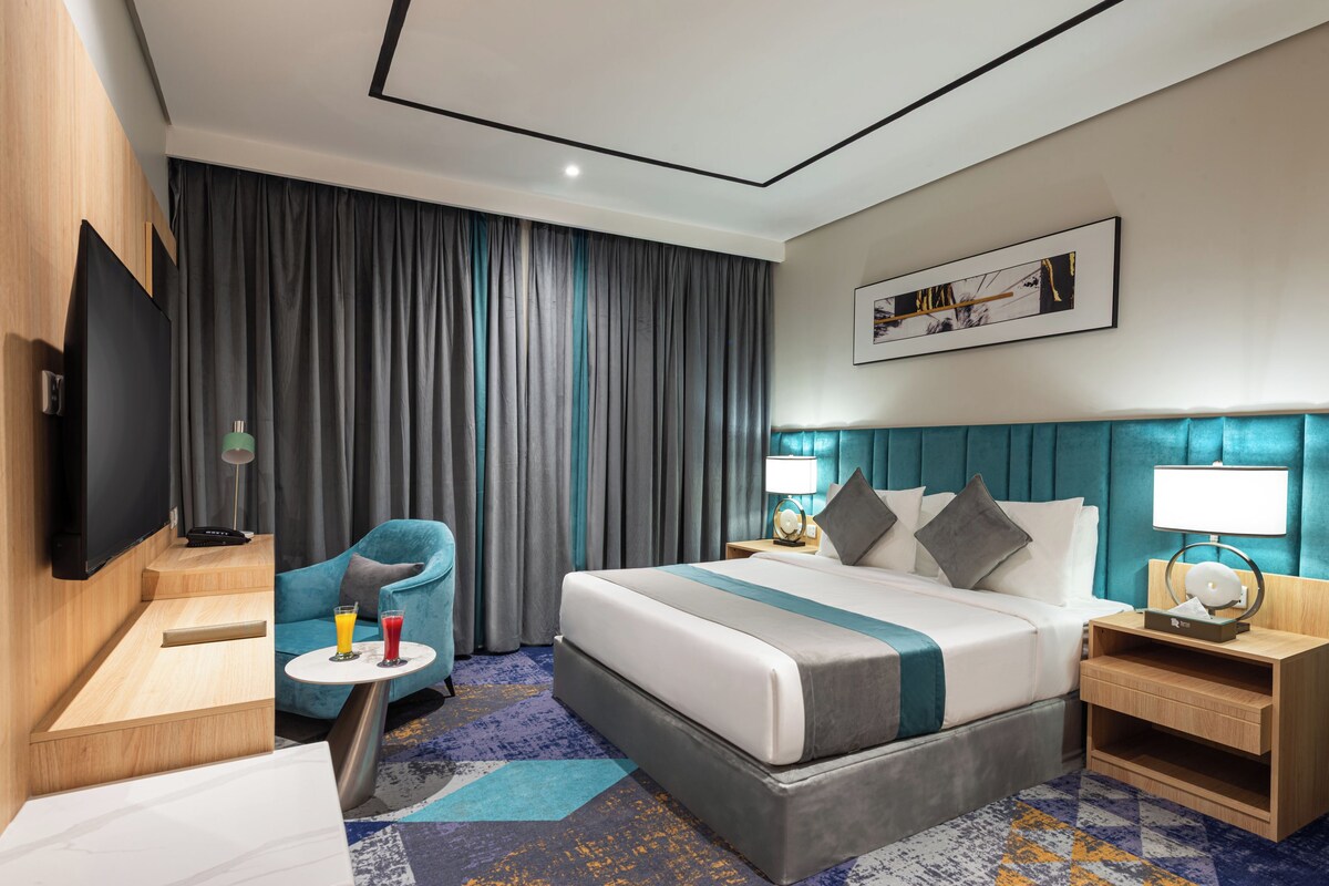 A hotel room with a bed, a TV, a side table, a chair, and a large window with curtains.