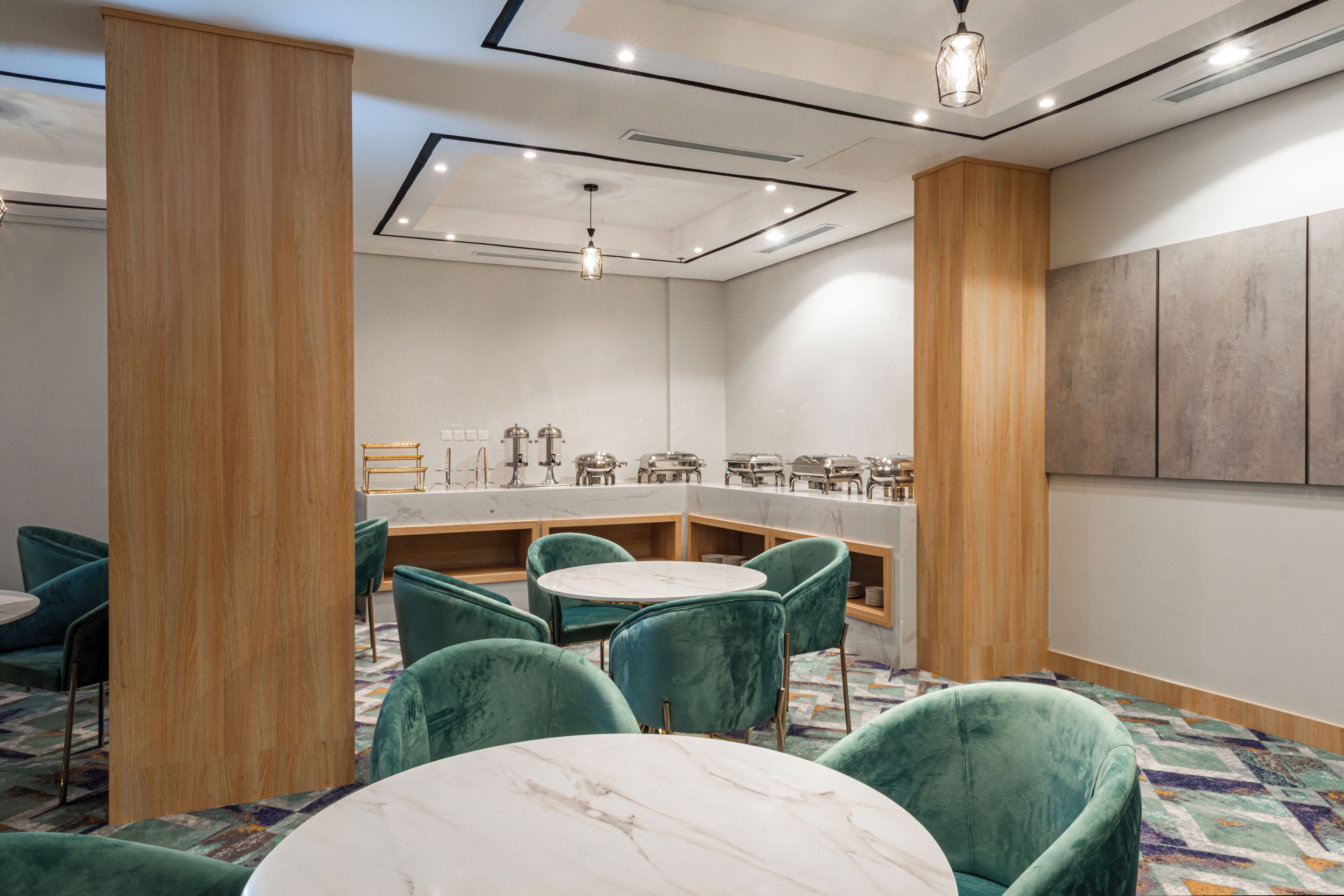 A modern hotel room with a marble-topped table, teal chairs, a wooden accent wall, and a built-in minibar.