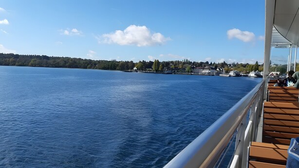 Blick vom Borddeck eines Schiffes mit Geländer, einem klaren, blauen Himmel und einer fernen Küstenlinie mit Gebäuden und Bäumen.