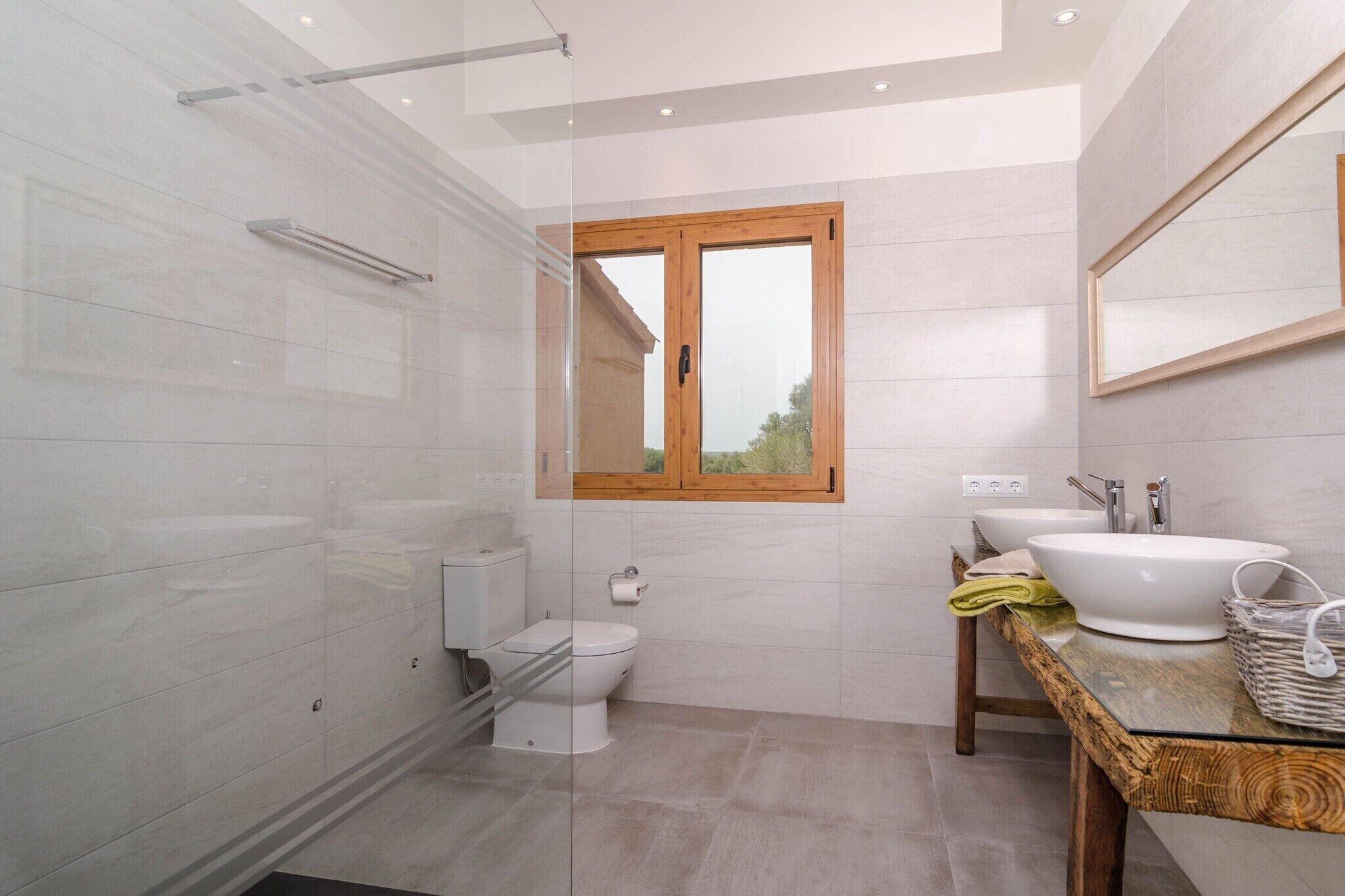 A modern bathroom with a glass shower enclosure, a toilet, a sink with a wooden countertop, and a window with a wooden frame.
