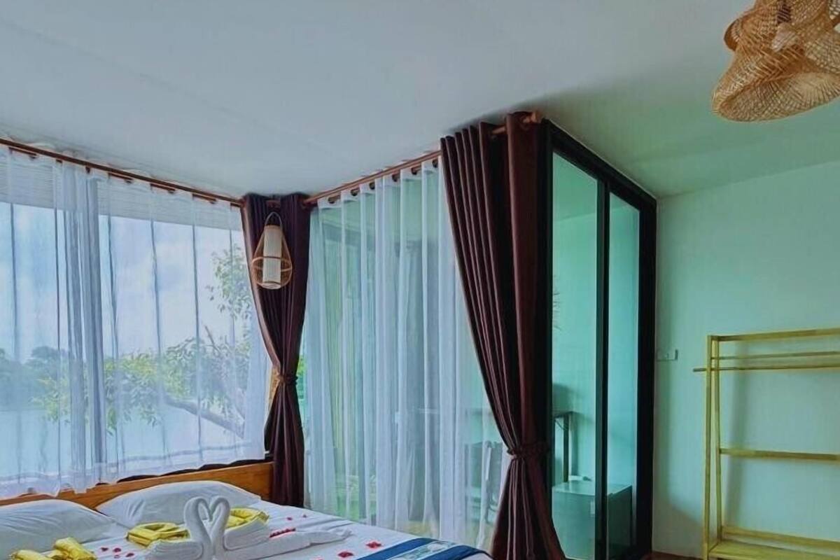 A hotel room with a bed, a window with sheer curtains, a wooden floor, and a balcony with a metal railing.