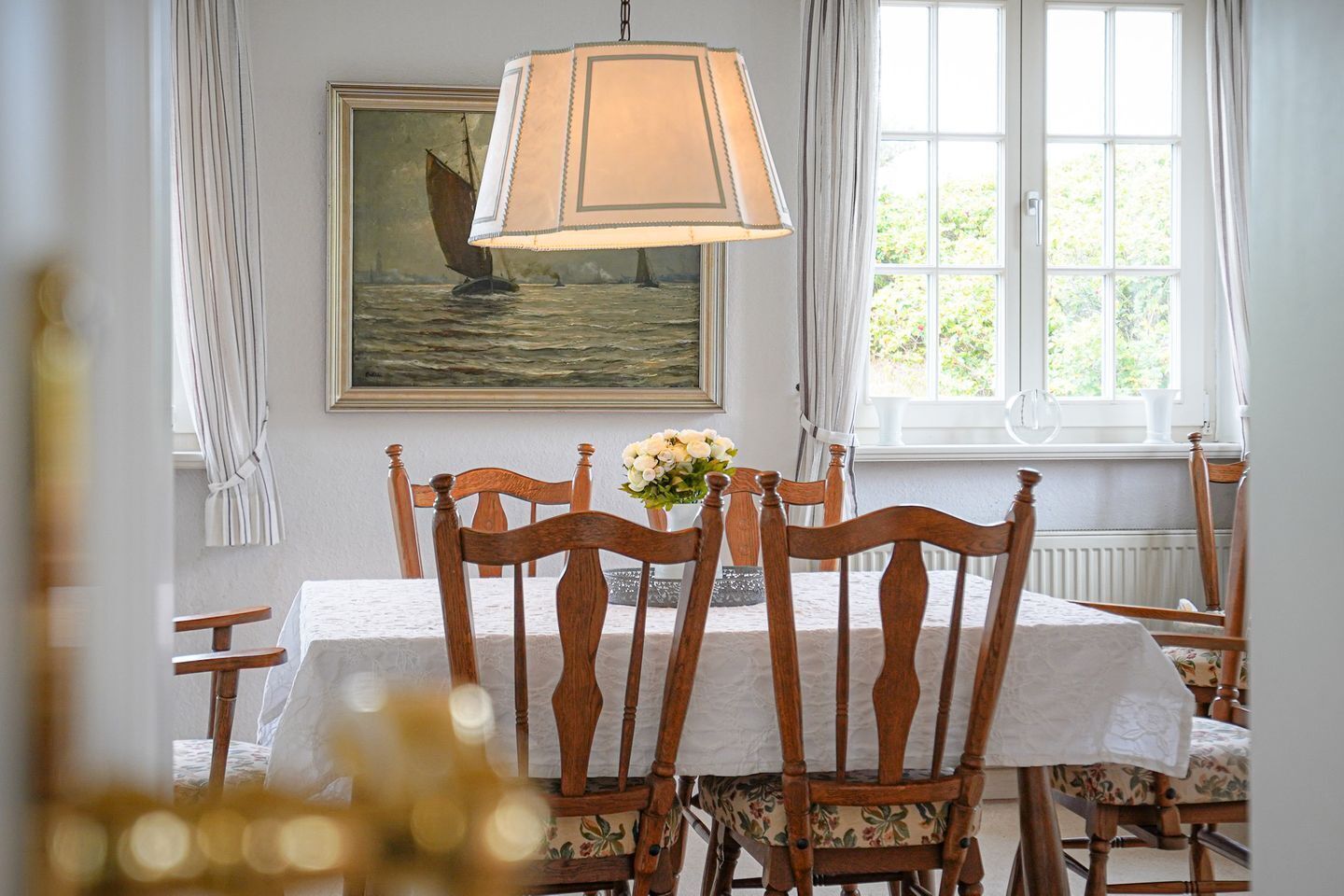 Un comedor con sillas de madera, una mesa con mantel blanco, una lámpara colgante y un cuadro de un velero en la pared.
