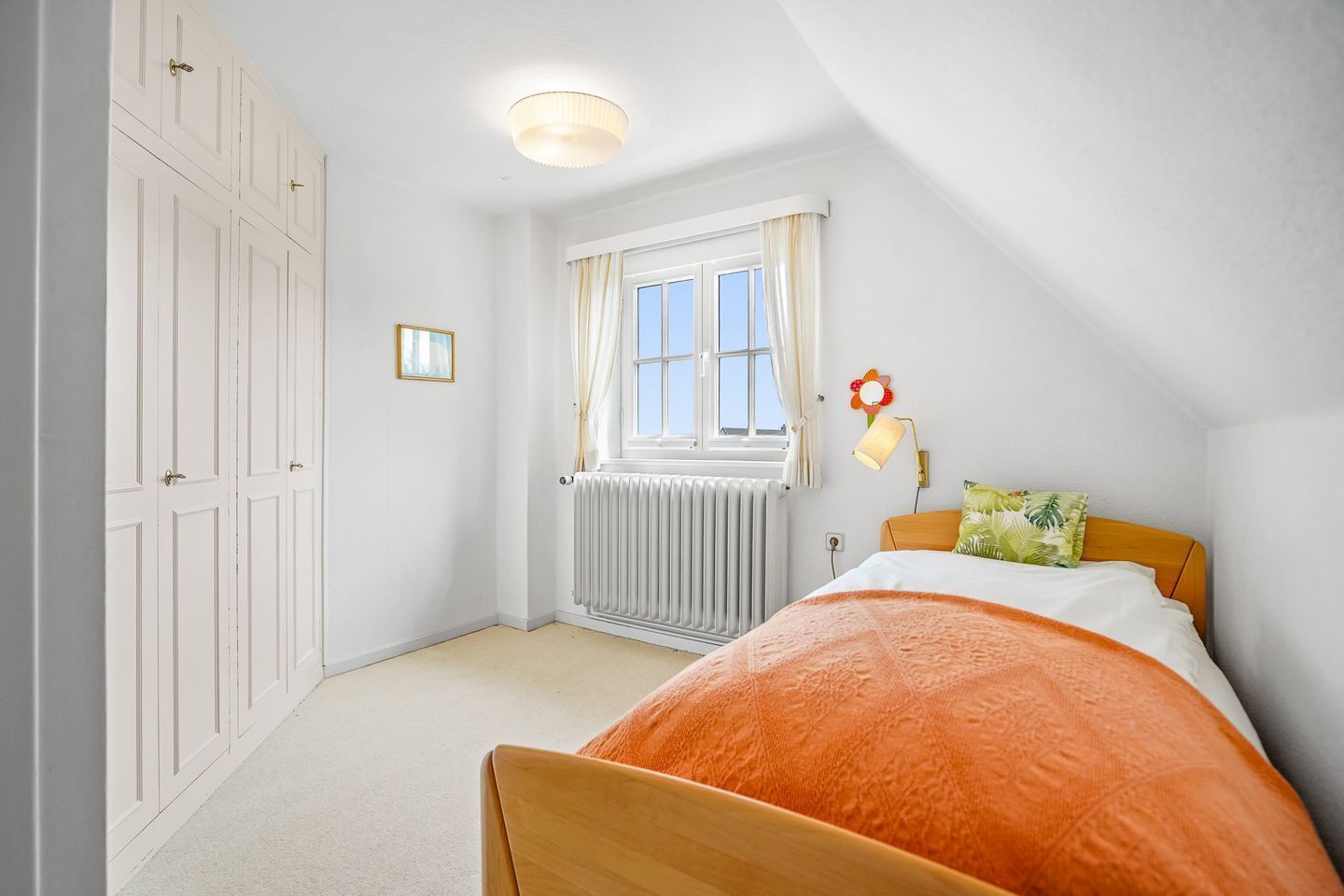 Une chambre à coucher comprenant un lit, un radiateur, une fenêtre avec des rideaux, une lampe fixée au mur et une porte.