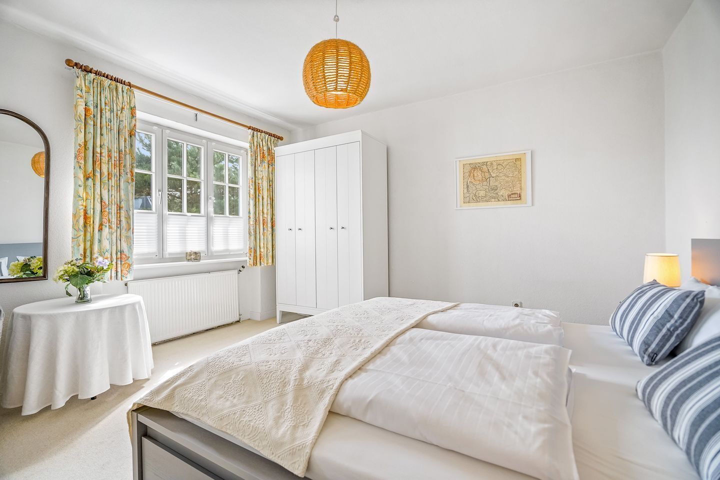 A bedroom with a bed, a bedside table, a lamp, a window with curtains, a radiator, and a hanging light fixture.