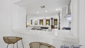 A modern kitchen with white countertops, stainless steel sinks, and a central island. There are two gold wire chairs in the foreground. - Reflections in the Sea by Wine Coast Holidays (ALDINGA BEACH)