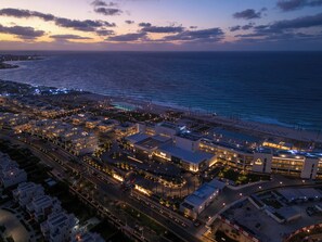 An aerial view of a coastal cityscape at dusk with illuminated buildings, a beach, and a marina. - Palace Beach Resort (El Alamein)