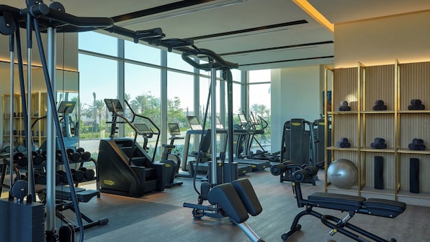 Une salle de sport moderne équipée de divers appareils d’exercice, dotée de grandes fenêtres et offrant une vue sur des palmiers.