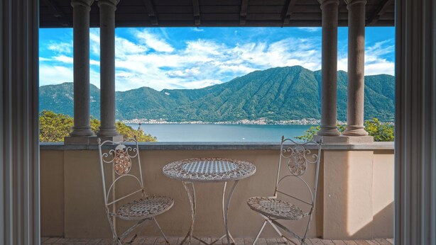 Un balcon avec deux chaises en métal et une petite table ronde, donnant sur un lac et des montagnes.