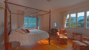 Un lit à baldaquin avec vue sur la mer, un plancher en bois et des fauteuils à motifs floraux.