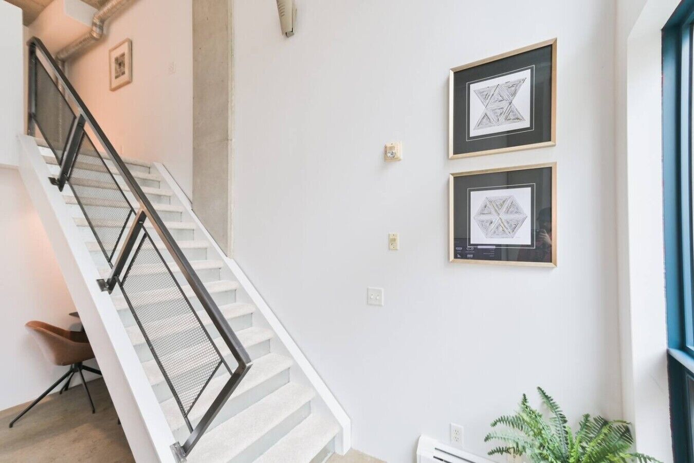 A staircase with a metal railing, a white wall with two framed artworks, and a plant near a window.