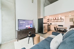 A living room with a flat-screen TV, a sofa, a coffee table, and a kitchen area with a refrigerator, microwave, and cabinets. - Trendy Loft | Downtown | UG Park | Sofa Bed (Calgary)