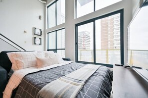 A modern bedroom with a large bed, multiple windows, and a view of a cityscape. - Trendy Loft | Downtown | UG Park | Sofa Bed (Calgary)