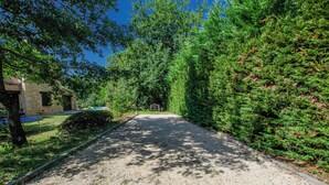 Un camino de grava conduce a una casa de fachada de piedra, rodeada de abundante vegetación y árboles.