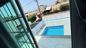 A view from a balcony with a swimming pool, a clear sky, and buildings in the background.