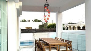 A modern outdoor dining area with a wooden table and chairs, a hanging decorative light fixture, and a view of a building in the background.
