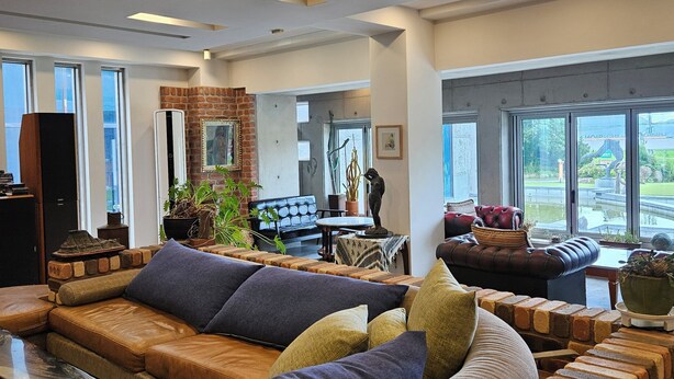 Uma sala de estar moderna com sofá seccional, parede de tijolos decorativa e grandes janelas com vista para o exterior.