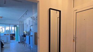 A hallway with a large mirror, a door, and a bench. There is a view into another room with a kitchen area.